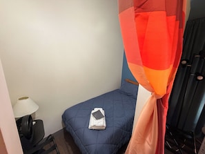 Una habitación pequeña con una cama individual, una mesita de noche, una lámpara y una cortina.