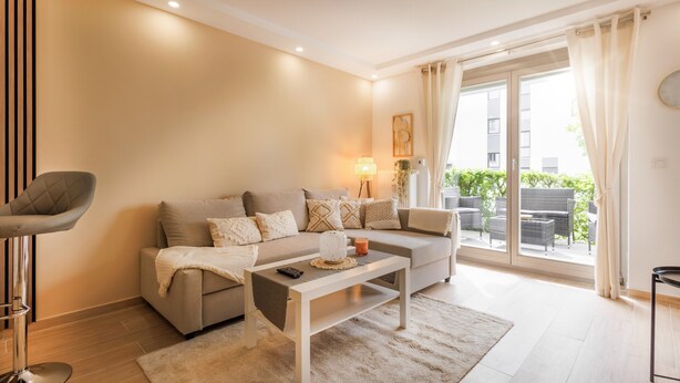 Ein modernes Wohnzimmer mit einer Couch, einem Couchtisch und einem Sessel. Ein Fenster mit Vorhängen, eine Lampe und Blick ins Grüne sind zu sehen.