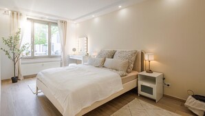 Ein Schlafzimmer mit einem großen Bett, Nachttischen, einem Spiegel, einer Zimmerpflanze und einem Fenster mit Vorhängen.