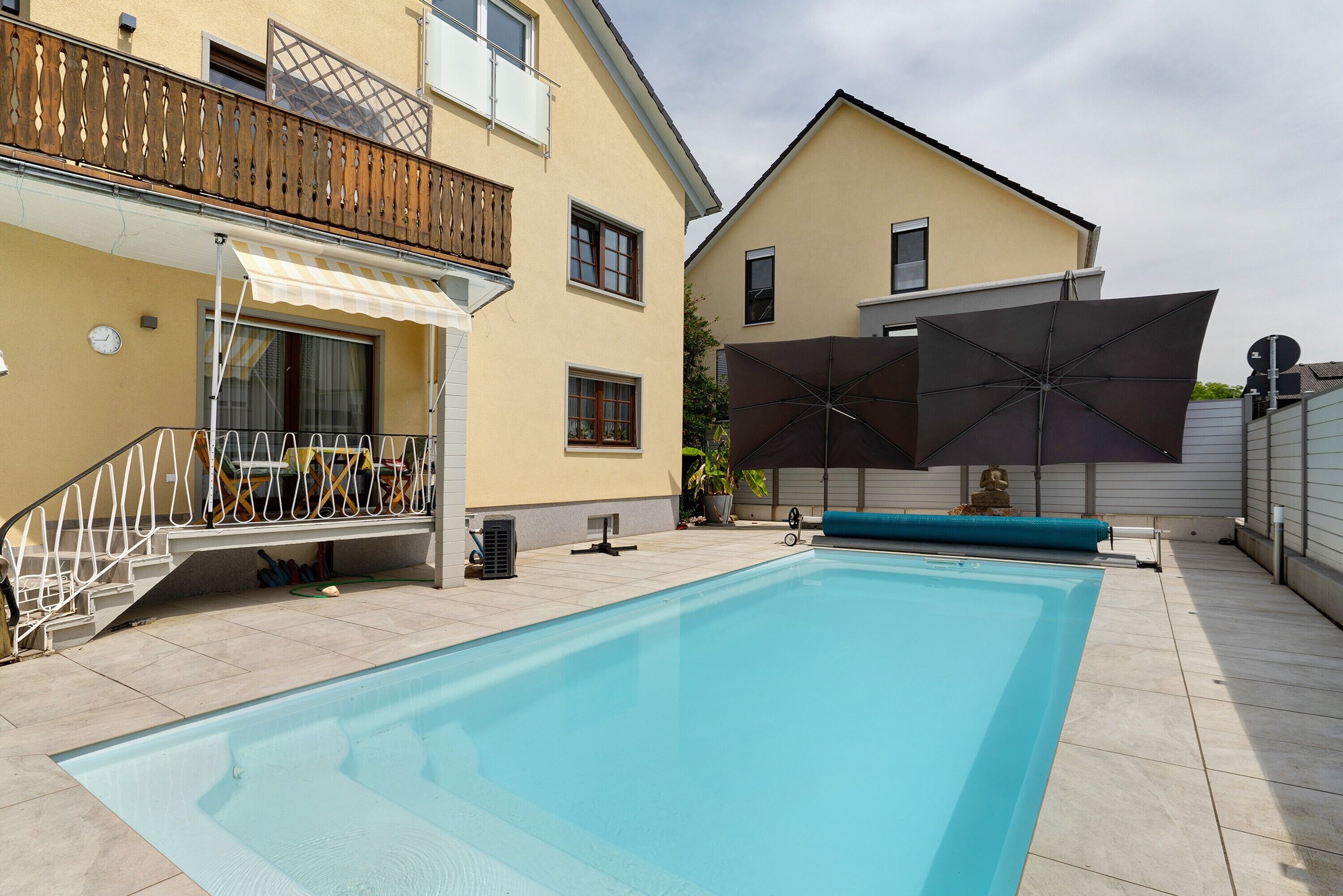 Ein Schwimmbecken mit Holzterrasse, einer überdachten Sitzgelegenheit und einem Gebäude mit Balkon.