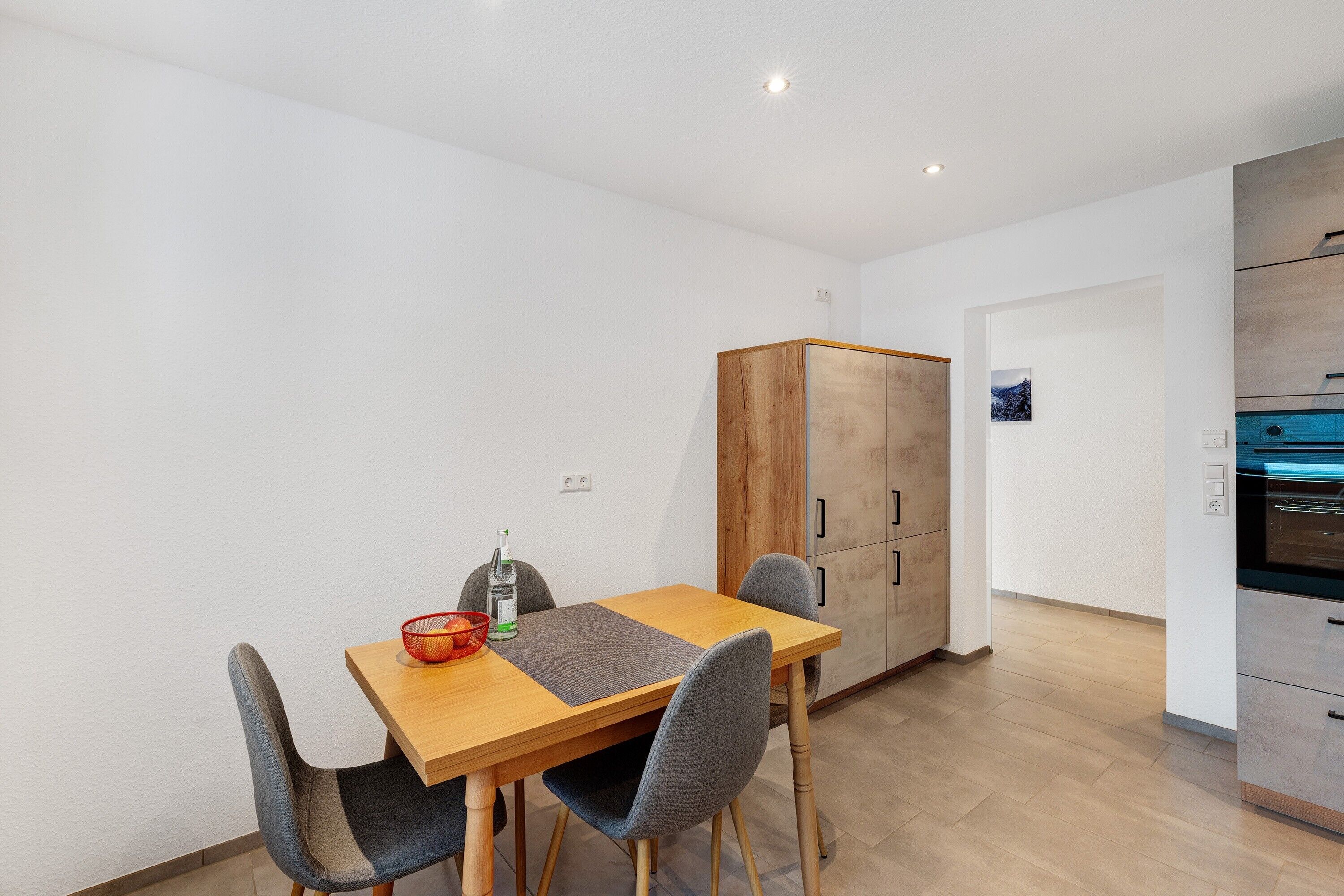 Een eethoek met een houten tafel, grijze stoelen, een rode schaal en een fles.