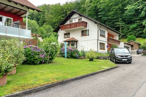 Ein zweistöckiges Haus mit Balkon, einem geparkten Auto davor und einem üppigen, grünen Garten.