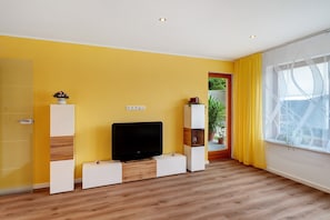 Ein Zimmer mit gelber Wand, einem Flachbildfernseher auf einem Holzboden, einer Topfpflanze und einem Fenster mit transparenten Vorhängen.