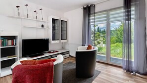 Un salon moderne doté d’un téléviseur à écran plat, d’étagères blanches, d’un pouf noir et d’une grande fenêtre agrémentée de rideaux.