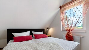Une chambre à coucher avec un lit, des oreillers, une table de chevet, une lampe, une fenêtre avec des rideaux et un radiateur.