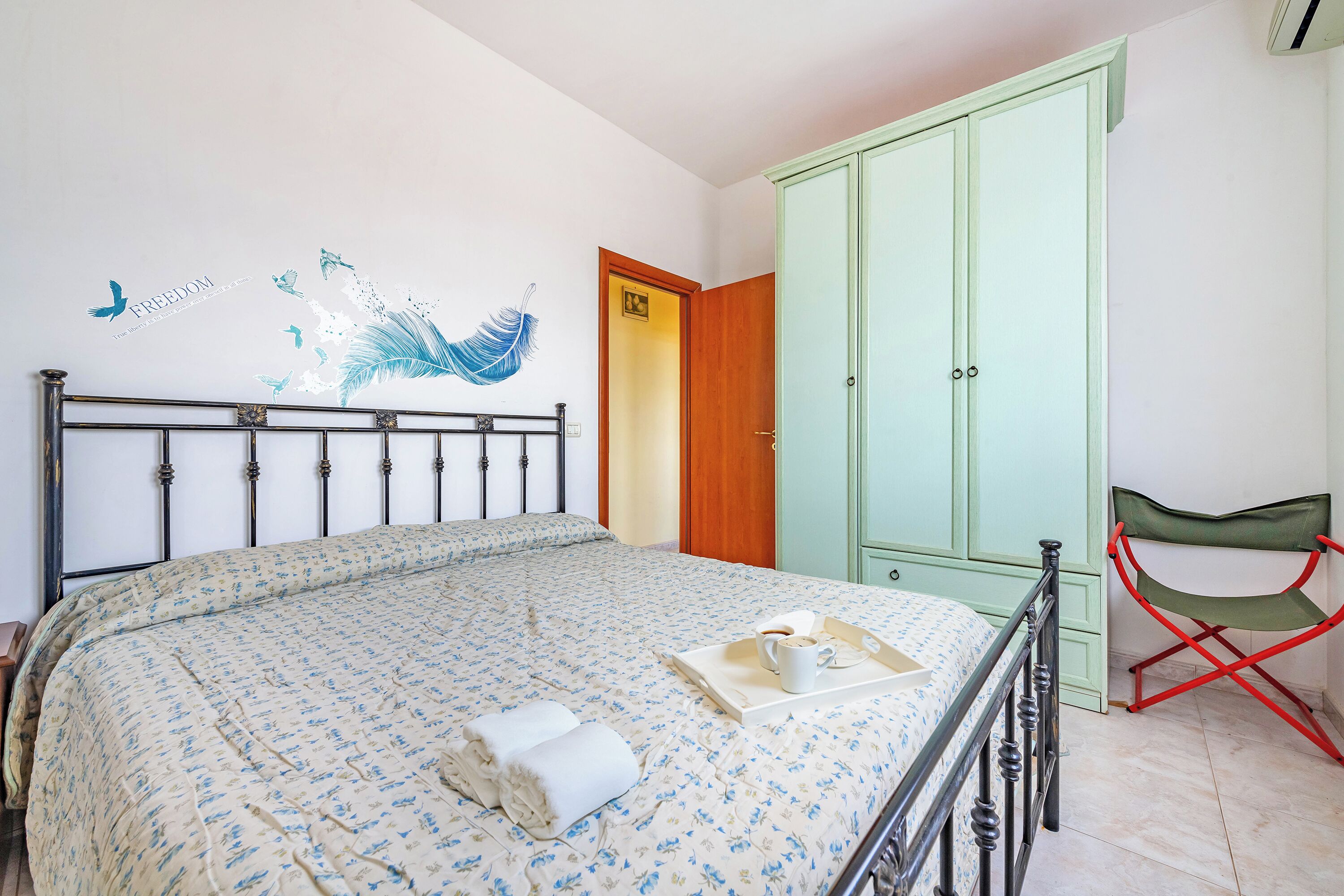 Une chambre à coucher avec un lit, une armoire verte, une chaise pliante rouge et un motif décoratif sur le mur.