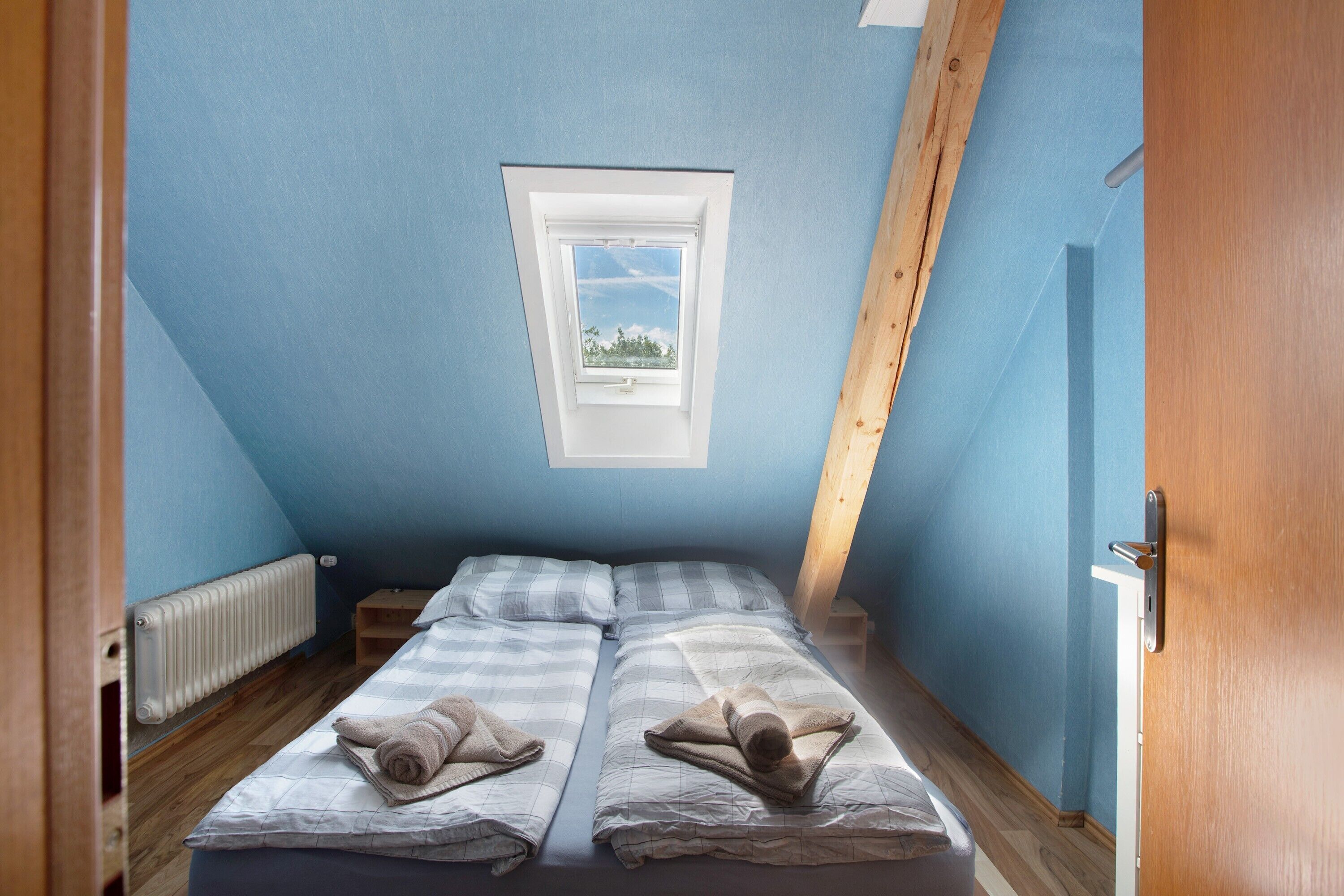A small, single-room with a sloped ceiling, a skylight, a radiator, and two beds with towels.