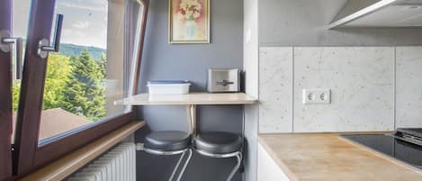 A kitchen with a window, a radiator, two stools, a shelf, and a painting.