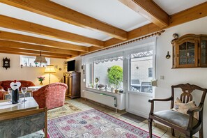 Un salon chaleureux avec des poutres en bois, une table à manger, un fauteuil rouge, un meuble en bois et une vue à travers une porte vitrée.
