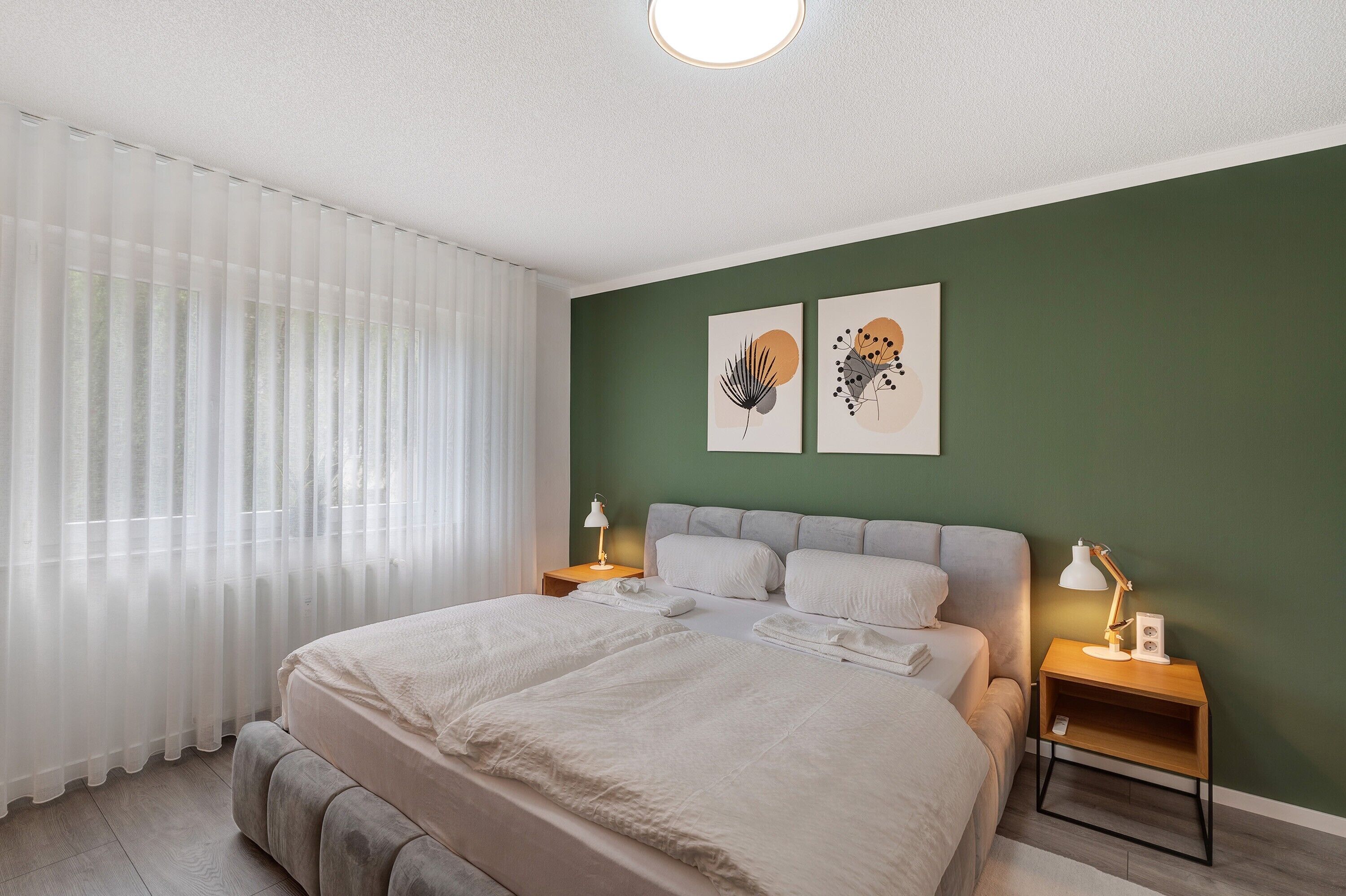 Ein Schlafzimmer mit einem Bett, zwei Nachttischen, einem Fenster mit transparenten Vorhängen und zwei Wandbildern.
