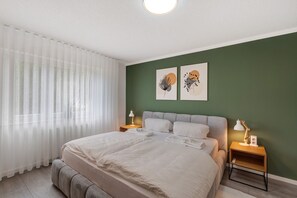 A bedroom with a bed, two bedside tables, a nightstand, a window with sheer curtains, and two wall art pieces.