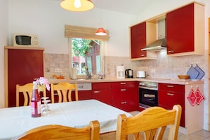 Een keuken met rode kastjes, een wit aanrecht, een eettafel met stoelen, een magnetron, een koffiezetapparaat en een raam met uitzicht op groen.