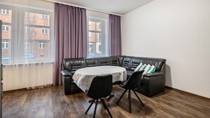 Um quarto com sofá de couro preto, mesa redonda com toalha de mesa estampada, duas cadeiras pretas, janela com cortinas leves e vista para prédios lá fora.