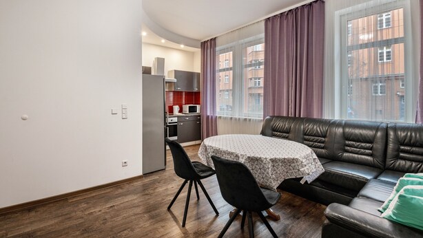 Uma sala de estar moderna com um sofá de couro, uma mesa de jantar com cadeiras, uma cozinha ao fundo e grandes janelas com cortinas.