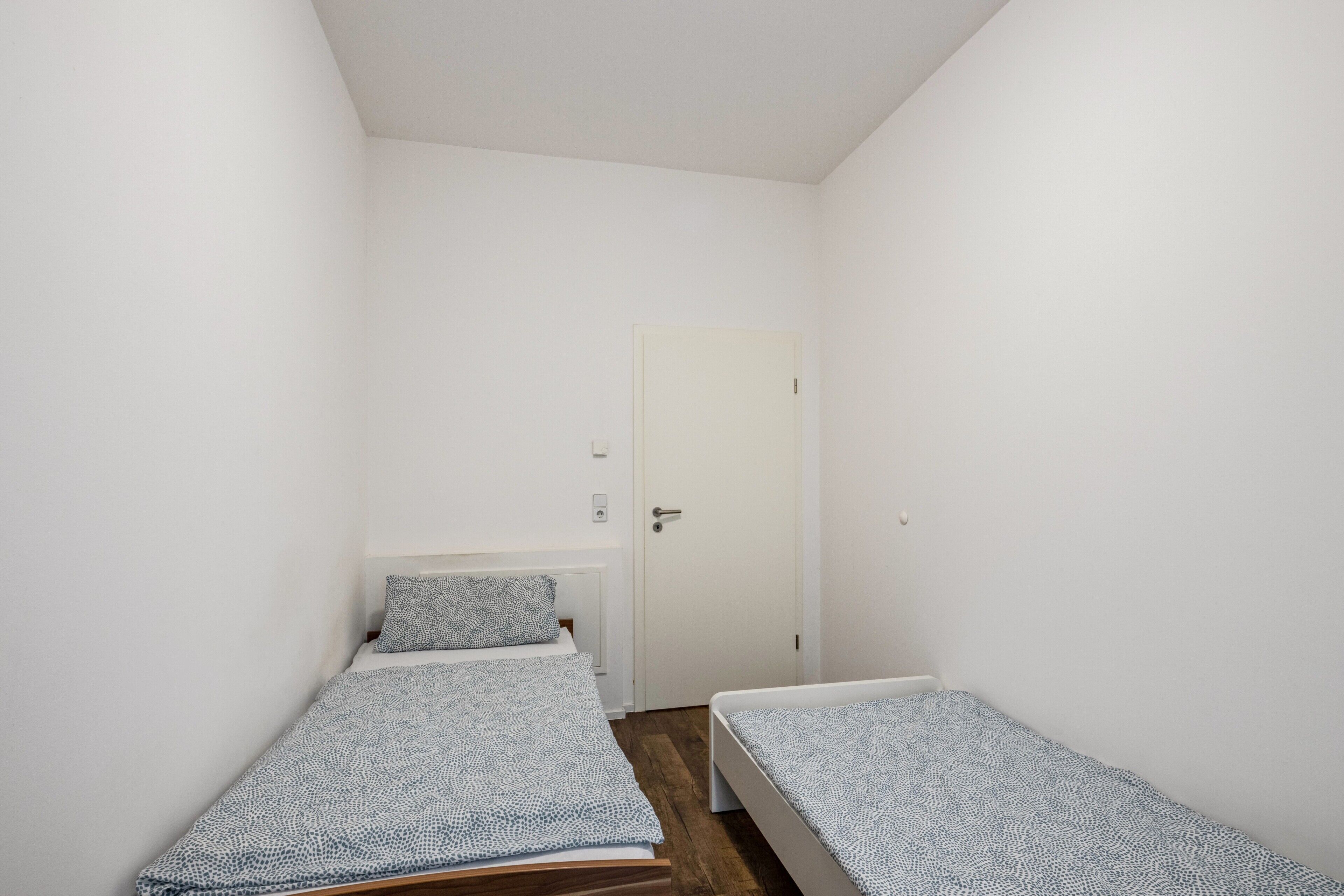 A small, narrow room with two single beds, a white door, and a white wall.