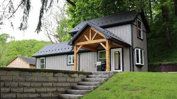 Une maison en bois à deux étages, dotée d’un mur de soutènement en pierre et d’un escalier en pierre menant à l’entrée.