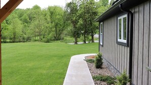 Une maison avec une façade en bois, un chemin en béton menant à la porte d’entrée et une pelouse verdoyante avec des arbres en arrière-plan.