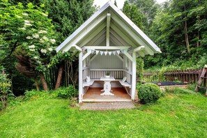A wooden gazebo with a bench and a table, surrounded by lush greenery.