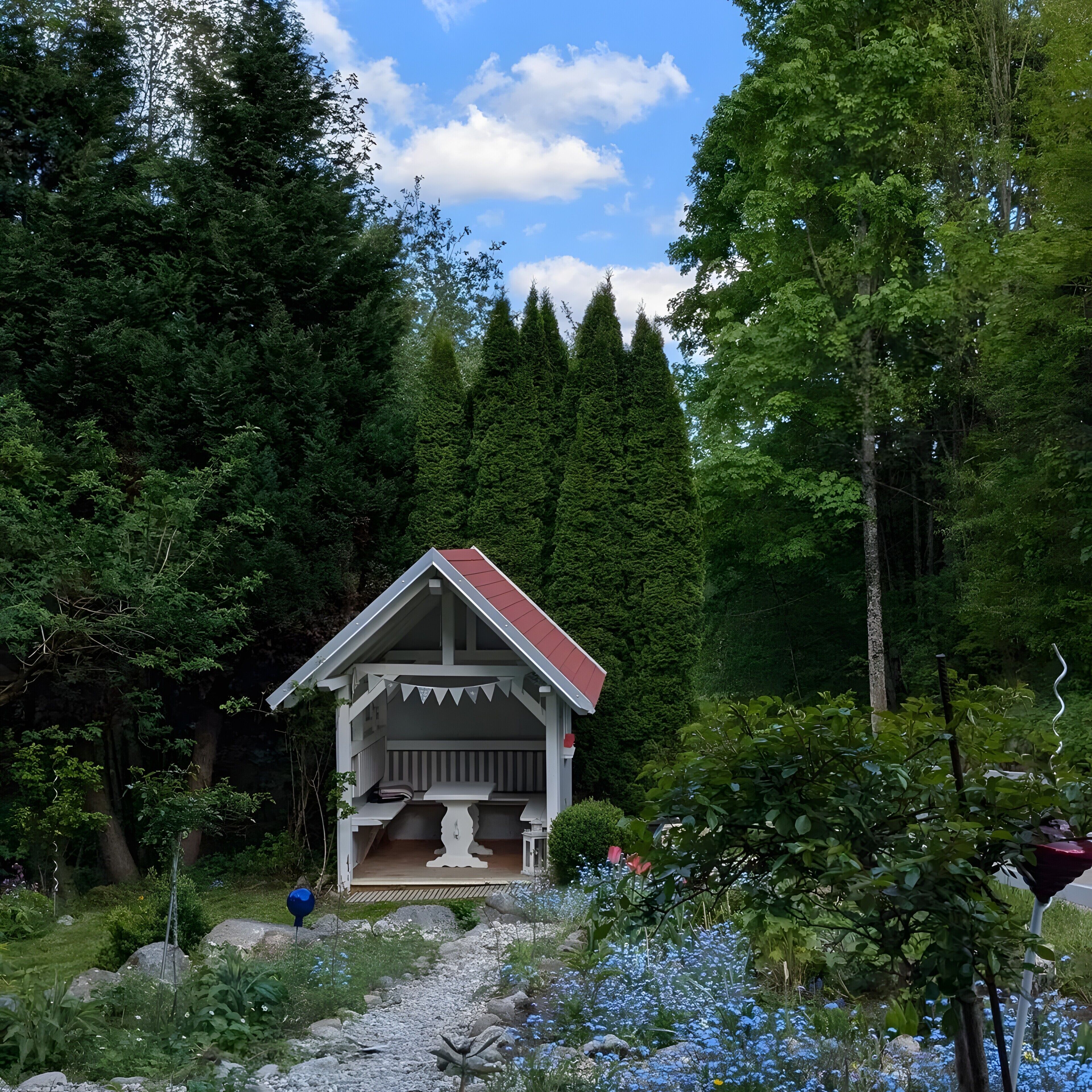 A small cabin with a red roof and white walls, surrounded by dense forest and a gravel path.