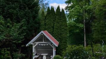 A small cabin with a red roof and white walls, surrounded by dense forest and a gravel path.