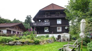 Een tweelaags huis met een balkon, een tuin met rotsen en planten, en een houten stoel.