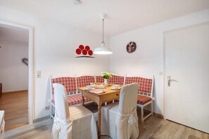 Un espace repas comprenant une table en bois, des chaises blanches, un banc à carreaux rouges et blancs, un luminaire suspendu blanc et un mur orné d’un motif rouge et noir.
