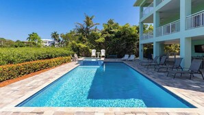 A rectangular swimming pool with clear blue water, surrounded by a tiled deck. There are lounge chairs and a small table with chairs on one side, and a building with balconies on the other.