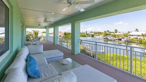 A covered outdoor seating area with white sofas and a table, overlooking a marina with boats and palm trees.
