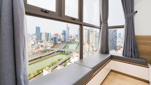 A room with a large window overlooking a cityscape, including a river and various buildings. - KaLa RiverGate Apartment - Ben Thanh (Ho Chi Minh City)