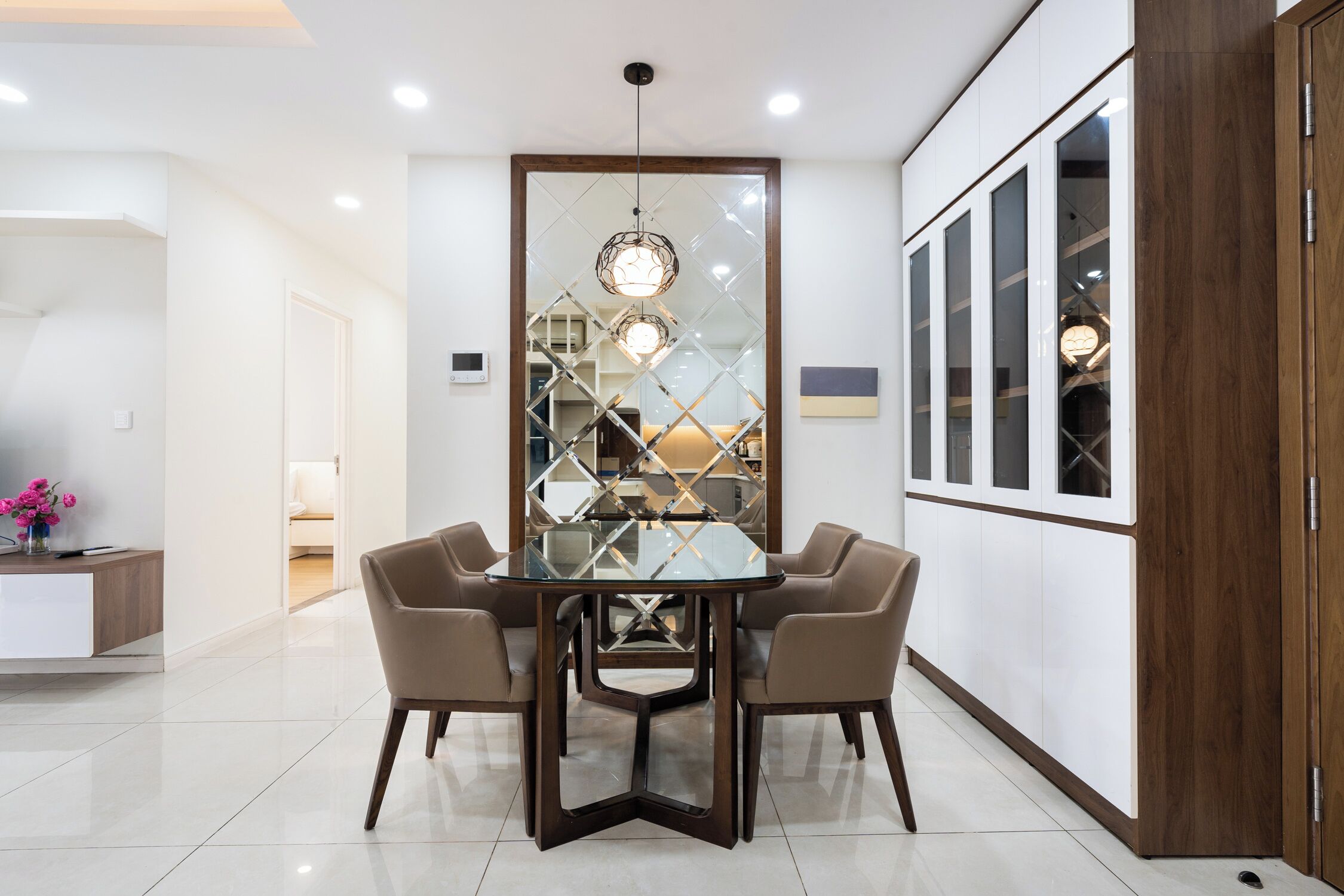 A modern dining area with a glass table and four chairs, a wooden cabinet with glass doors, and a decorative wall panel.