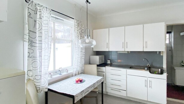 A compact kitchen with white cabinets, a small dining table, and a window with patterned curtains. - 9 Person Holiday Home in Sovik (Ålesund)