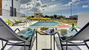 Un espace piscine extérieur moderne, doté de chaises longues, d’une table avec une assiette de nourriture et offrant une vue sur les bâtiments environnants.