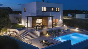 Une maison moderne à deux étages, dotée d’une piscine, d’un espace extérieur aménagé et d’un patio bien éclairé.