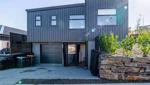 A modern two-story building with a garage door, a small entrance, and a stone wall with plants. - Cosy Kirimoko Escape in Wanaka (Wānaka)