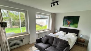A bedroom with a large window, a grey sofa, a bed with white linens, a radiator, and a small table.