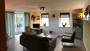 Un soggiorno con divano, tavolino, televisione e libreria. Sono presenti finestre con tende e una pianta sospesa al soffitto.