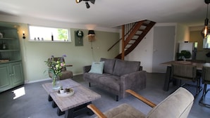 Un soggiorno accogliente con un divano grigio, un tavolino in legno e una poltrona beige. Una scala conduce a una zona cucina con un tavolo e sedie in legno.