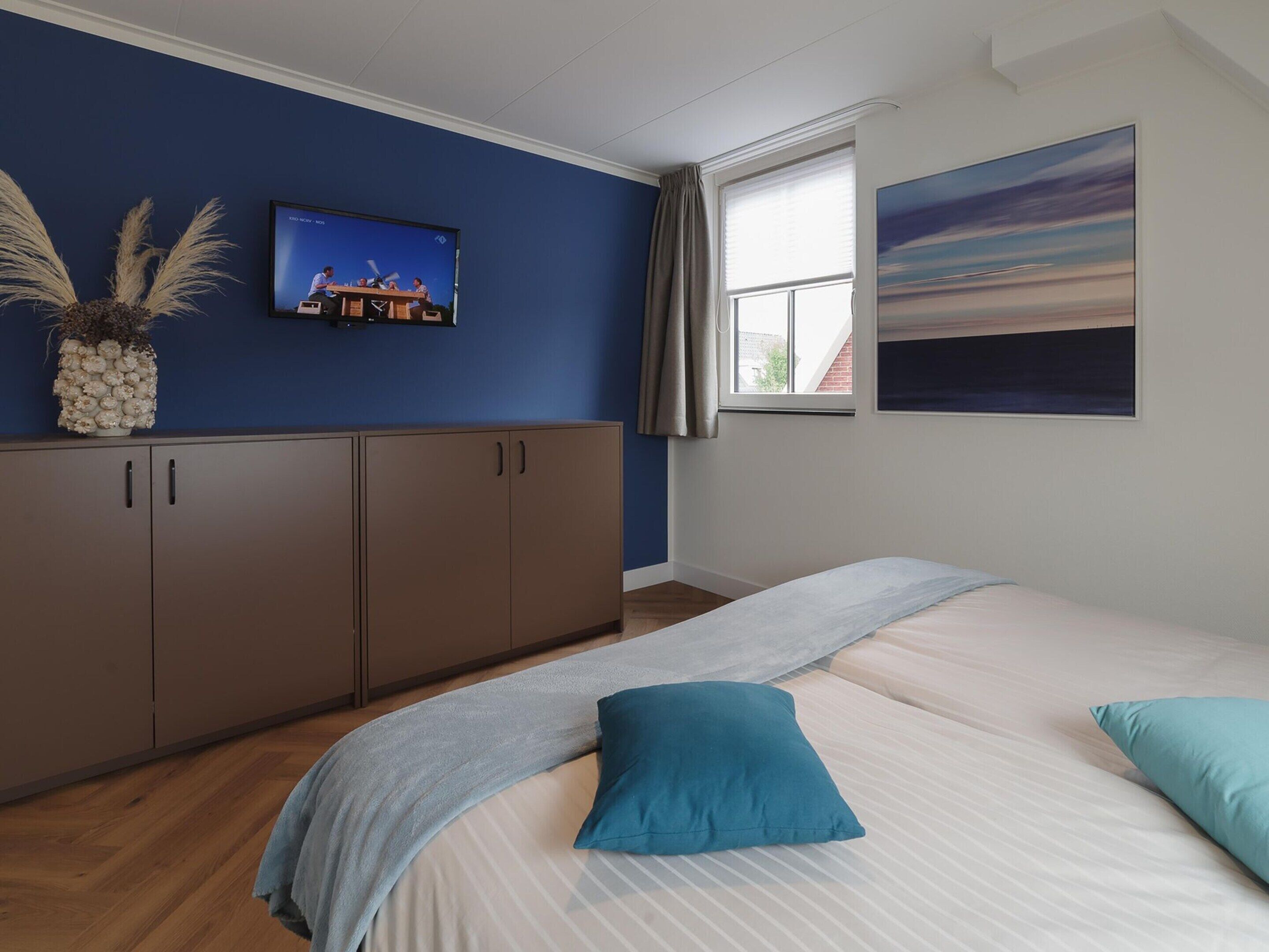 A bedroom with a bed, a wardrobe, a window with curtains, a painting, and a television on the wall.