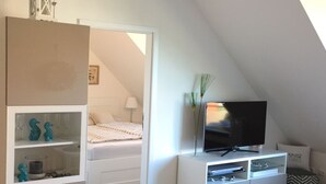 Une chambre à coucher avec un lit, un téléviseur posé sur un meuble blanc, une petite étagère avec des objets décoratifs et une fenêtre laissant entrer la lumière naturelle.