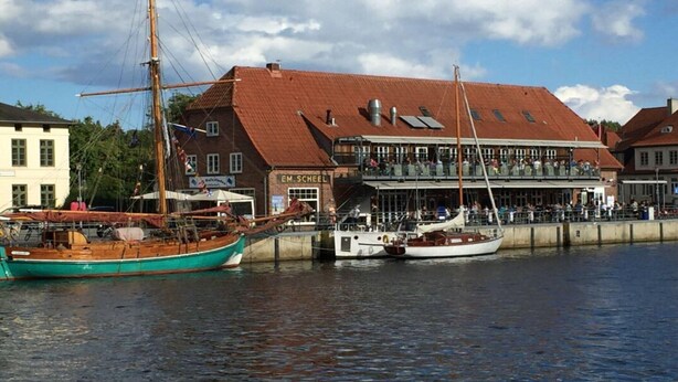 Une promenade en bord de mer, avec un bâtiment traditionnel doté d’un toit rouge et d’une enseigne indiquant « EM. Scheel ».