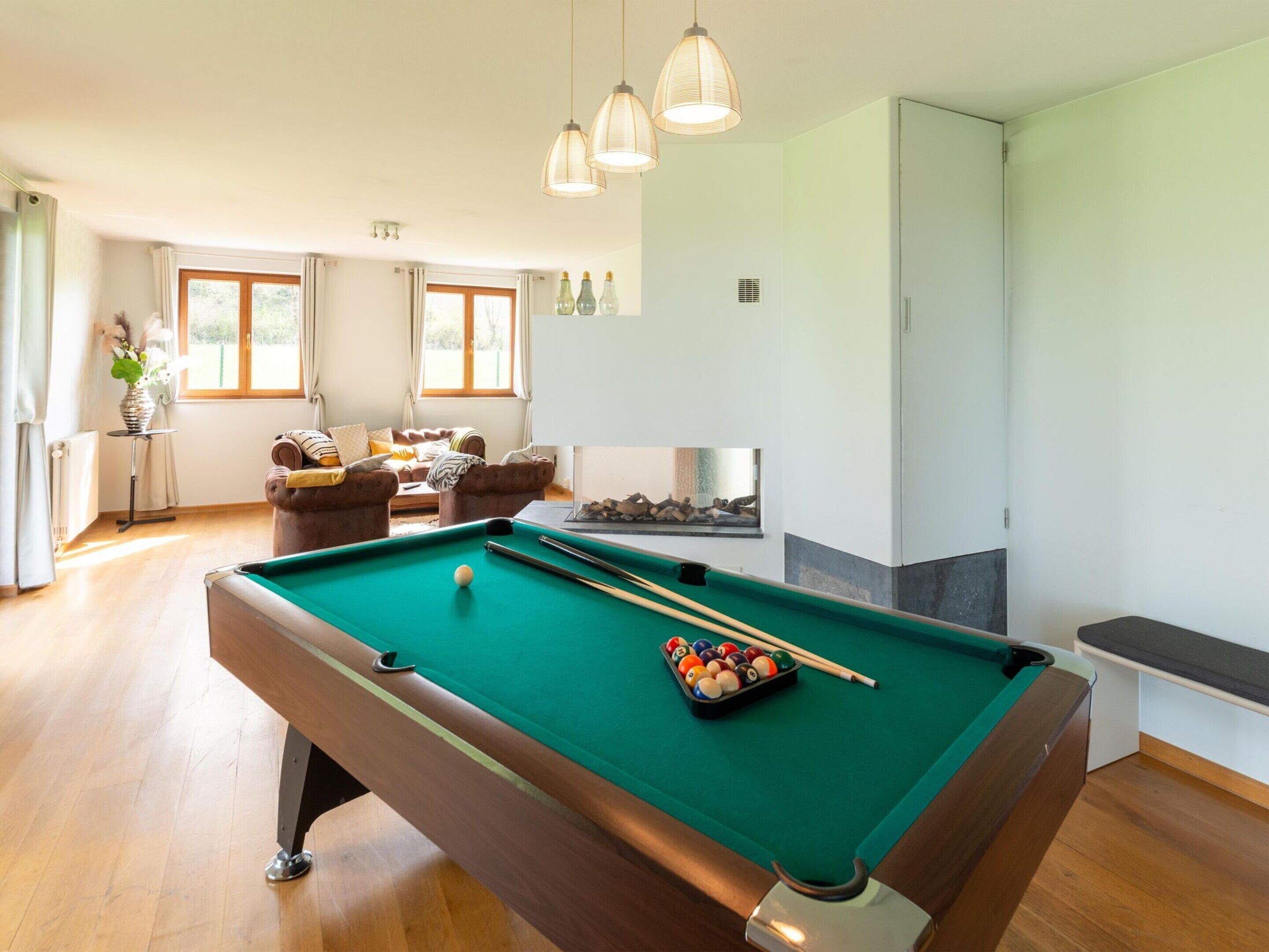 Une table de billard dans une pièce avec un canapé, une cheminée et de grandes fenêtres.