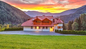 A two-story chalet with a red-tiled roof, set against a mountainous backdrop during sunset.