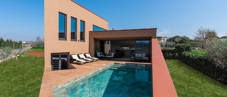 A modern house with a swimming pool, a patio with lounge chairs, and a clear blue sky.