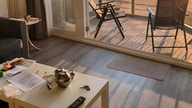 A room with a wooden floor, a sofa, a table with a remote control, and a view of the beach through a glass door. - Helmduin 5D Comfortable Holiday Residence (Egmond aan Zee)