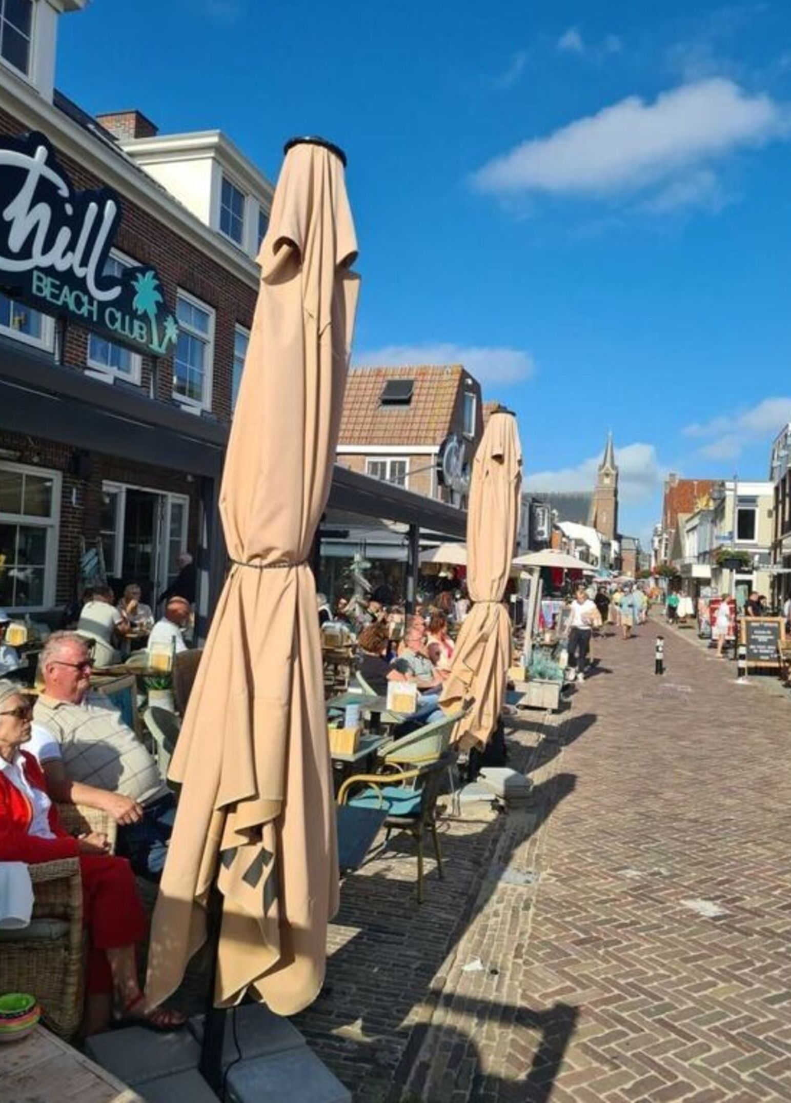 A bustling street with outdoor seating covered by beige umbrellas, a visible beach club sign, and a brick building with a tower.