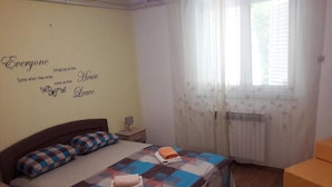 Une chambre à coucher comprenant un lit, une table de chevet, une fenêtre avec des rideaux légers et un mur orné d’une inscription décorative.