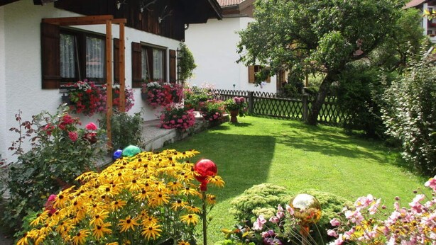 A white building with a sloped roof and wooden shutters, surrounded by a well-maintained garden with various flowers and a lawn. - Holiday Apartment Maurer Apartment Laber (Oberammergau)