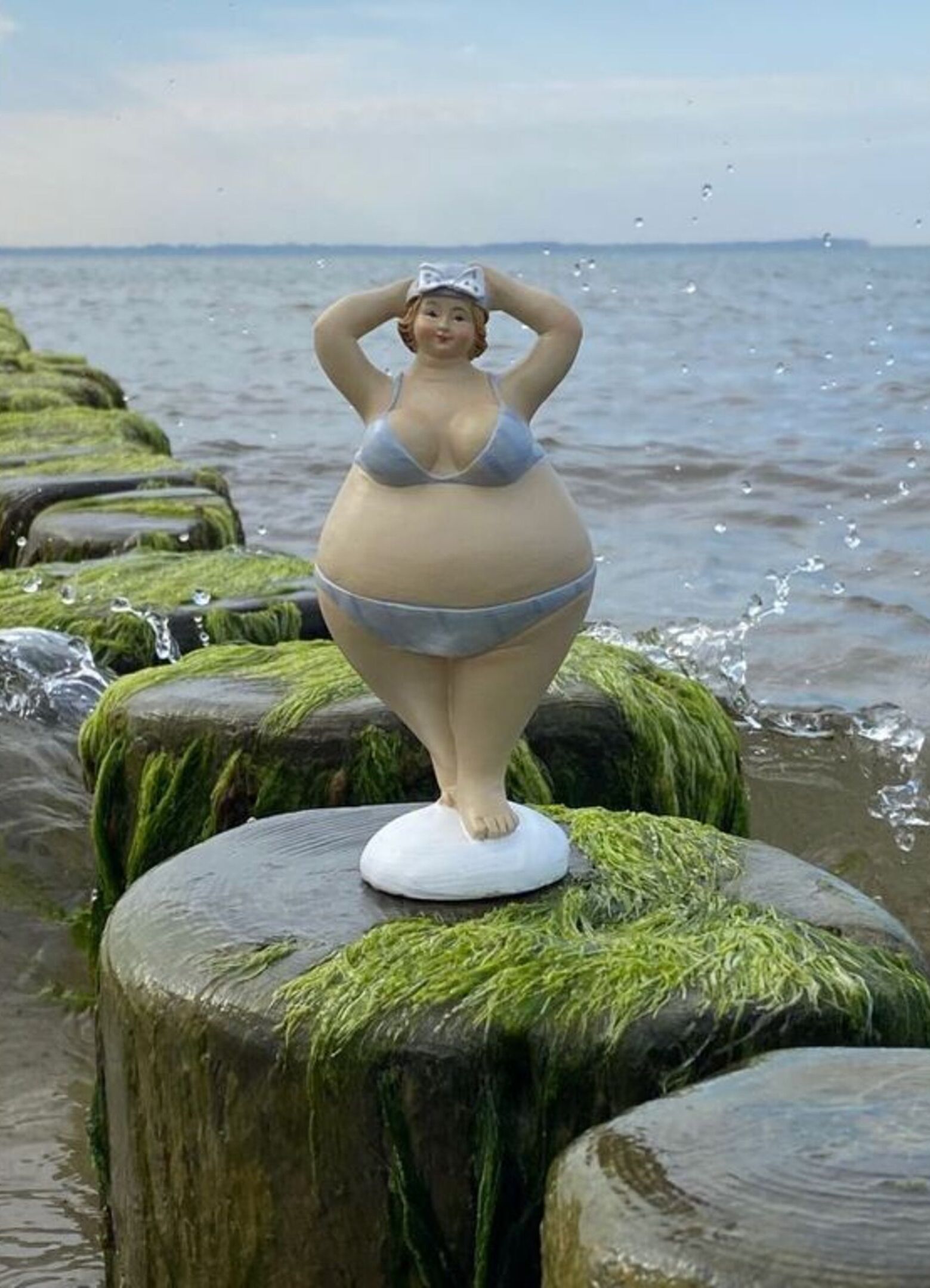 A statue of a woman in a bikini on a moss-covered post by the sea.