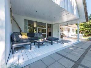 Uma área de estar moderna ao ar livre, com um sofá e duas mesinhas de centro, um alpendre coberto e vista para o interior da casa.