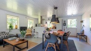 A spacious living area with a dining table, chairs, and a sofa. There is a kitchen area with a stove and cabinets. The room has large windows and a ceiling with wooden beams. - 4 Star Holiday Home in Norre Nebel (Norre Nebel)